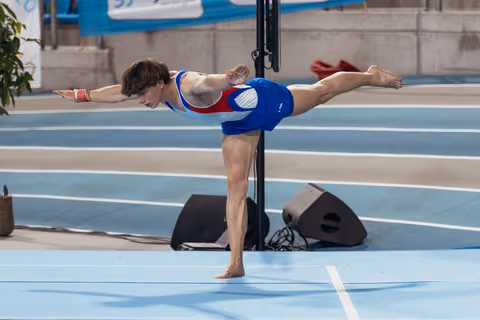 Young male gymnast holds a perfect arabesque balance on one leg, arms extended, focused expression, on blue competition floor.
