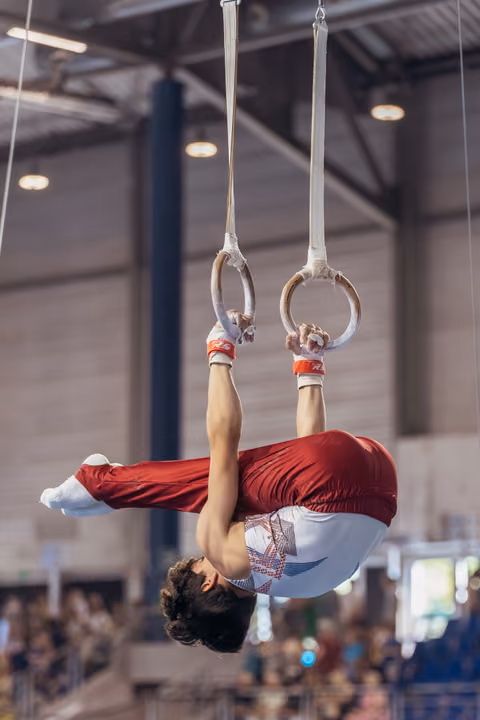 Young male gymnast performs an inverted hang on still rings, body fully extended, in a packed gymnastics hall.