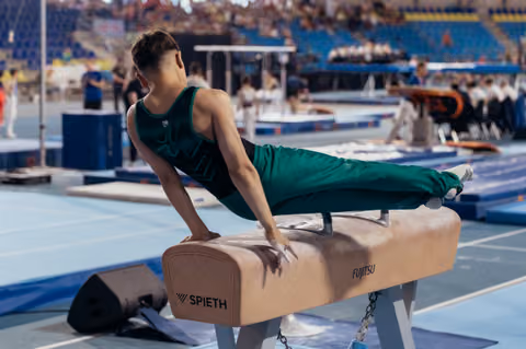 Young male gymnast in green leotard performs a straddle hold on the pommel horse during a competition.