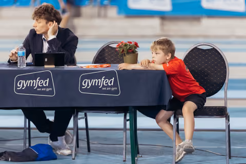A young boy in red leans restlessly on a Gymfed judges' table while an official works quietly beside him.
