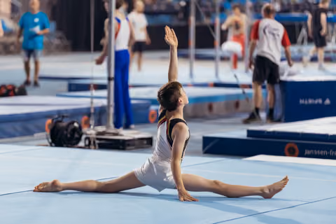 Young male gymnast performs a split on the floor exercise mat, one arm raised gracefully, in a competition arena.