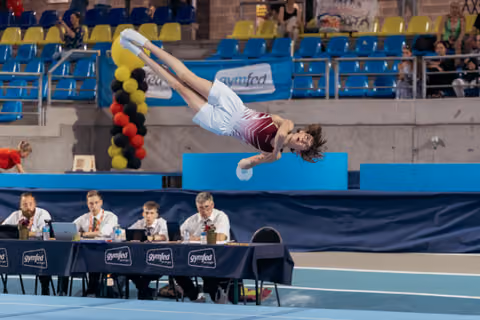 Young gymnast executes a powerful backflip mid-air during floor exercise at a Gymfed competition, judges seated below.