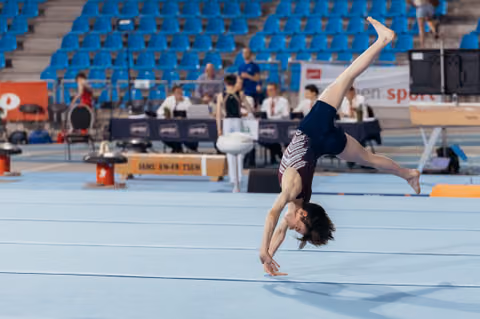 Young male gymnast performs a dynamic one-armed handstand on the floor exercise mat during a gymnastics competition.