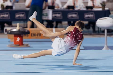 Young male gymnast performs a dramatic floor routine, balancing on one hand with legs split mid-air in competition.