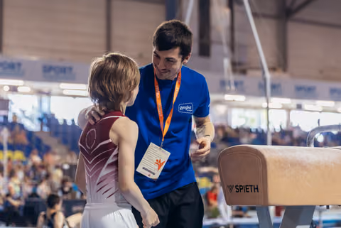 Smiling coach in blue Gymfed shirt places hand on gymnast's shoulder, offering encouragement beside a Spieth pommel horse.