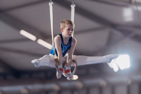 Young male gymnast holds a tuck position on still rings, wearing a blue leotard, with intense concentration.