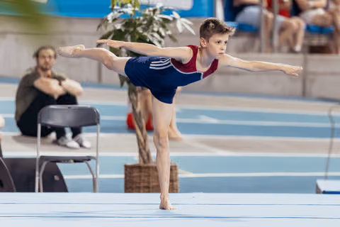 Young male gymnast holds a precise arabesque on floor, arms outstretched, intense focus on his face in an indoor arena.