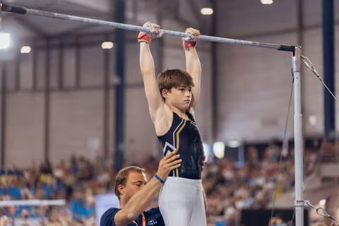 Young male gymnast hangs focused on high bar, coach steadying him from below in packed gymnasium.