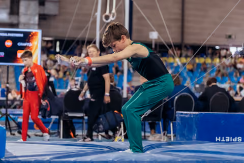 Young male gymnast in green uniform chalks his hands with intense focus before competing on rings at indoor meet.