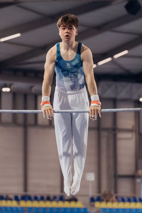 Young male gymnast grips the high bar with intense focus, body suspended mid-routine in an indoor gymnastics arena.