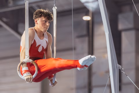 Young male gymnast performs on still rings, legs raised, wearing red uniform, expression of intense concentration.