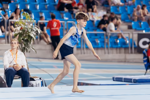 Young male gymnast runs barefoot across the floor exercise mat with focused concentration during a gymnastics meet.