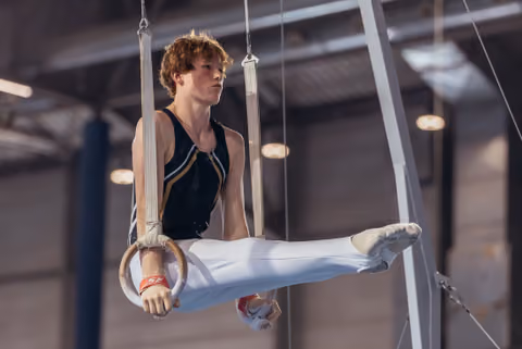 Young male gymnast performs an L-sit on still rings, expression focused and intense, in an indoor gymnasium.