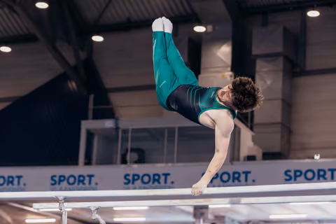 Male gymnast holds a dramatic inverted position on parallel bars, green leotard, intense focus, indoor arena.