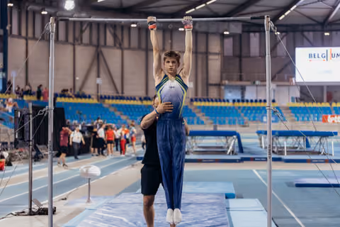 Young male gymnast hangs from high bar with focused expression while coach steadies him in Belgian sports hall.