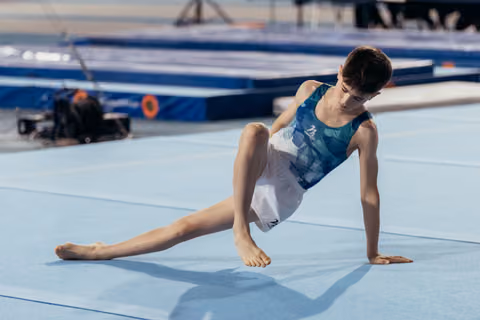 Young male gymnast stretches on a blue floor mat, concentrating intensely during warm-up at a gymnastics event.