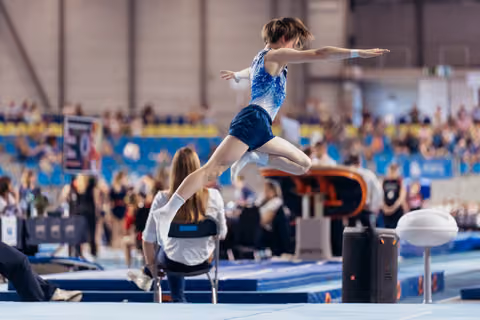 Young gymnast mid-leap on the floor exercise, arms outstretched, focused and composed during a competitive gymnastics event.