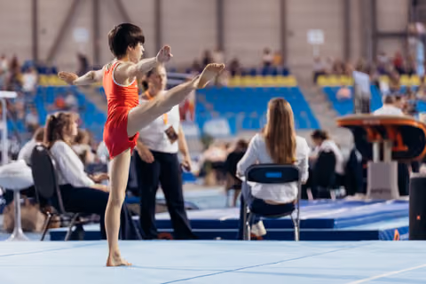 Young gymnast in red leotard performs a high leg raise on the floor exercise mat, focused and precise.