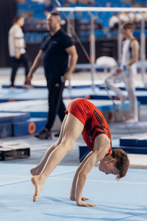 Young gymnast in red leotard performs a backbend on the blue floor mat, showing focus and flexibility.