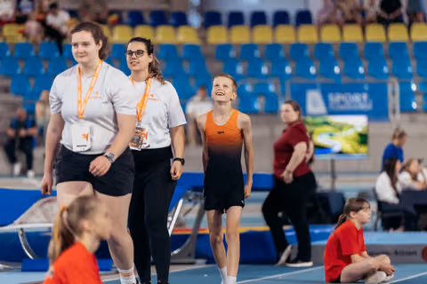 A smiling young gymnast in orange walks alongside two coaches in white shirts with lanyards on an indoor track.