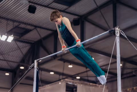 Focused young male gymnast grips the high bar with intensity, body extended, competing in an indoor gymnastics hall.