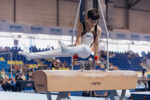 Young male gymnast performs on pommel horse, focused and controlled, wearing Belgian colors at an indoor sports arena.
