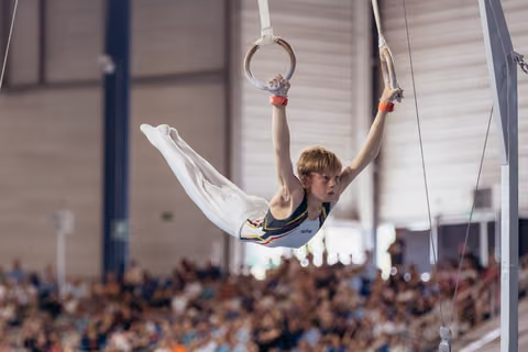 Young male gymnast holds a split position on still rings, focused expression, competing before a large indoor crowd.