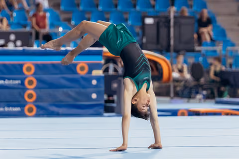 Young gymnast performs a backbend on the floor exercise mat during a competition, legs extended mid-movement.