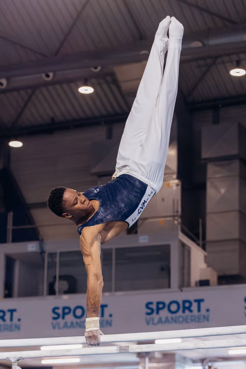 Male gymnast holds a powerful one-arm handstand on parallel bars, legs raised high, at Sport Vlaanderen event.