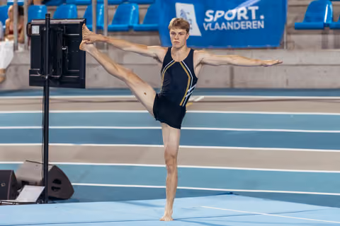 Young male gymnast holds a perfect standing split, arms outstretched, focused expression on blue indoor track.