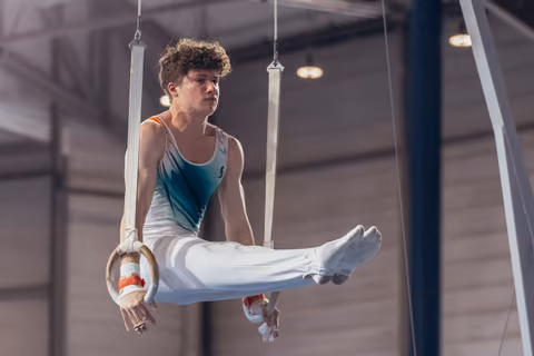Young male gymnast performs an L-sit on still rings, his focused gaze and taut muscles showing intense concentration.