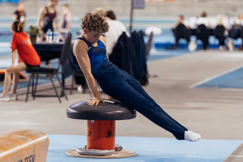 Young male gymnast performs a pommel mushroom routine, body extended, focused expression, at an indoor gymnastics competition.