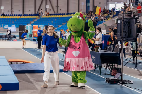 Smiling woman in blue shirt walks hand-in-hand with green GYM mascot waving Belgian flag at indoor gymnastics event.