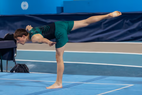 Gymnast in green leotard holds a focused arabesque pose on the blue floor exercise mat, arms extended forward.