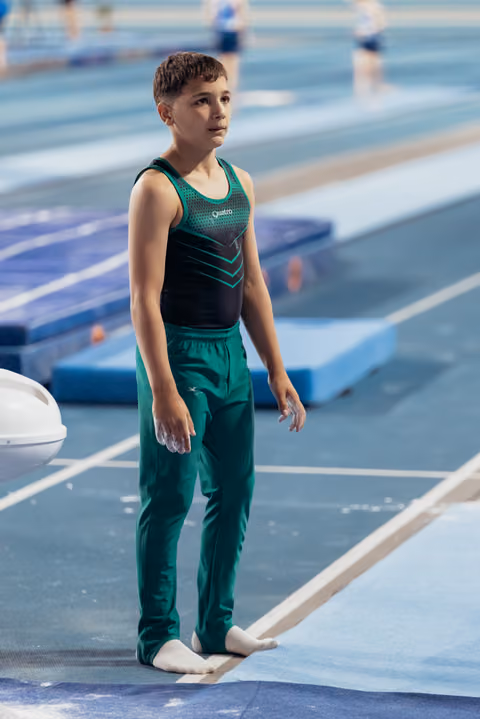 Young male gymnast stands barefoot on the floor exercise mat, looking up with focused concentration, wearing green and black attire.