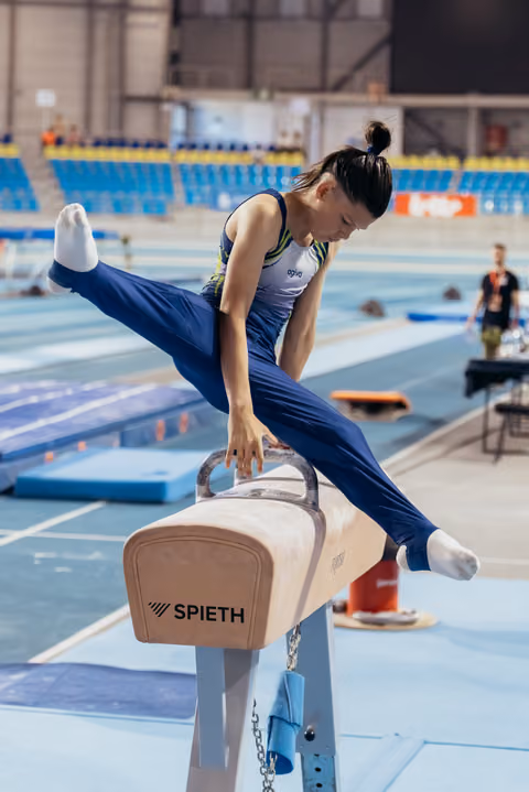 Female gymnast in blue leotard performs on a Spieth pommel horse, legs split wide, focused expression in a competition arena.