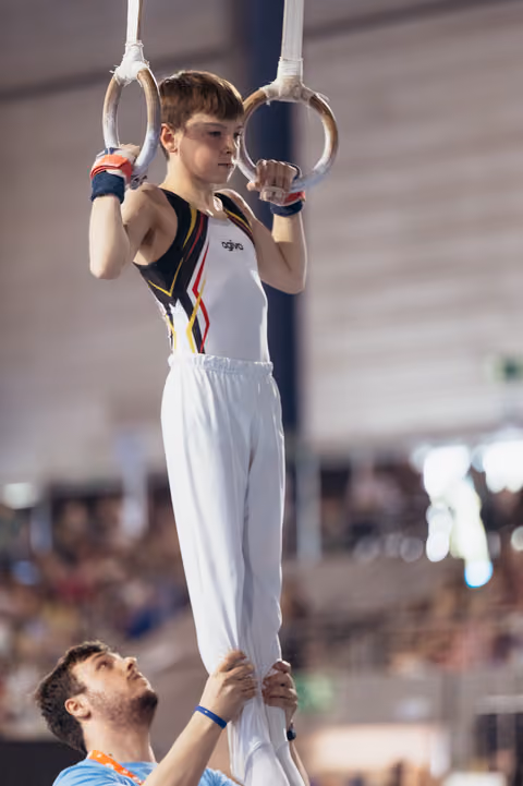 Focused young gymnast grips still rings with determination while coach steadies him from below in a packed arena.