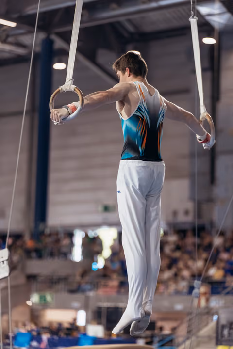Male gymnast performs on still rings, suspended mid-air in a teal leotard, showing controlled strength before a crowd.
