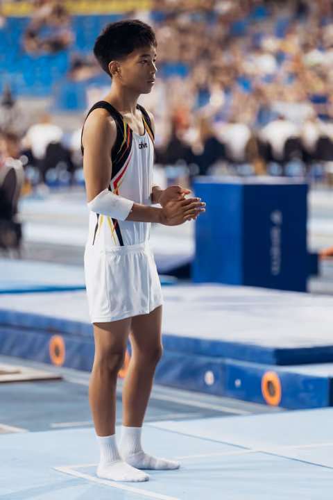 Young male gymnast stands poised, clapping hands together in concentration, wearing white uniform with black and red details in a packed arena.