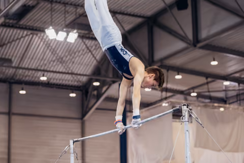 Young male gymnast holds a handstand on the horizontal bar, body fully extended, focused expression in an indoor gym.