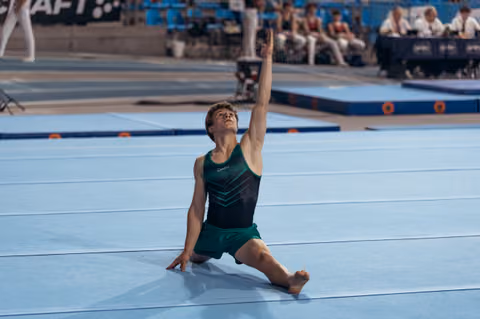 Young male gymnast in green leotard performs dramatic floor pose, arm raised skyward, head tilted back with intensity.