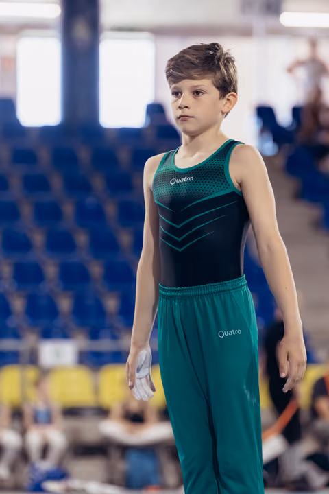 Young male gymnast stands still with focused expression, wearing Quatro leotard and green trousers, holding chalk-covered hand.