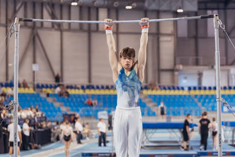 Young male gymnast hangs from the high bar with focused determination, wearing a teal leotard in a large competition arena.