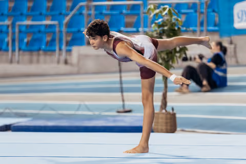 Young male gymnast holds a precise arabesque balance on the floor, focused expression, in a blue-seated arena.