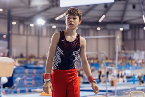 Young male gymnast stands composed on the floor, gazing upward with focused determination, wearing red wristbands.