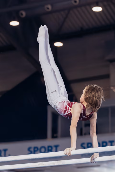 Young gymnast performs a precise handstand on parallel bars, legs extended upward, in a lit sports hall.