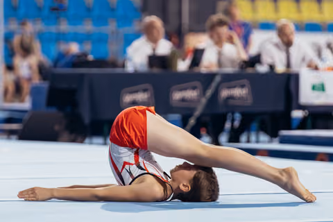 Young male gymnast performs an extreme backbend on the floor mat, judges watching in the background.