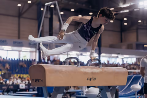 Young male gymnast performs on pommel horse, leaning forward with intense concentration in a packed arena.