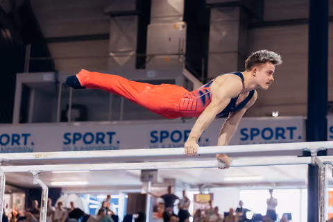 Male gymnast holds a planche on parallel bars, body horizontal, expression intense and determined at indoor sports event.