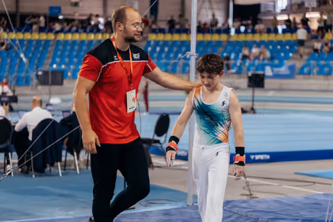 Coach places a reassuring hand on a young gymnast's shoulder as they walk away, the athlete looking downcast in a competition arena.